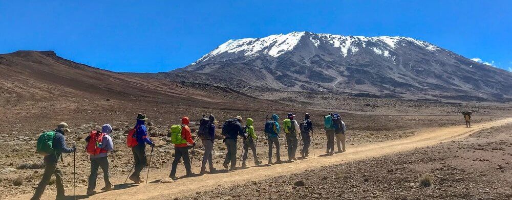 a group of people walking on a dirt path with a snowy mountain in the background